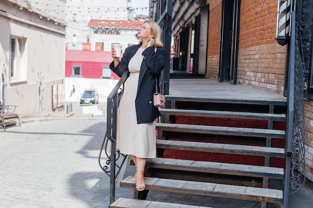 Beautiful young woman in dress and jacket walks around the city and drinks coffee