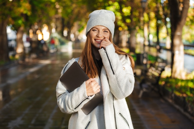 Beautiful young woman dressed in autumn coat and hat walking outdoors, carrying laptop computer