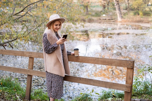 beautiful young woman drinking coffee, talking on phone in nature by the lake in autumn park in fall