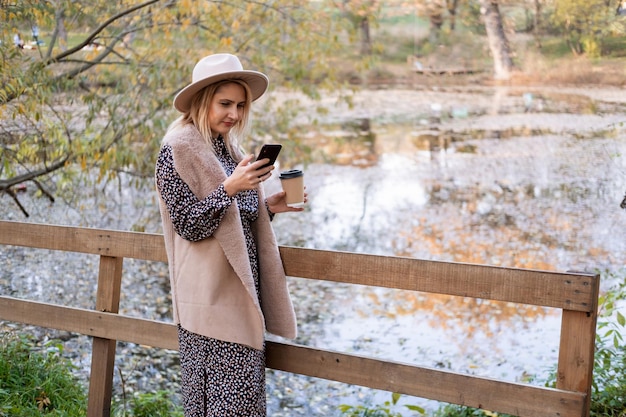 beautiful young woman drinking coffee, talking on phone in nature by the lake in autumn park in fall