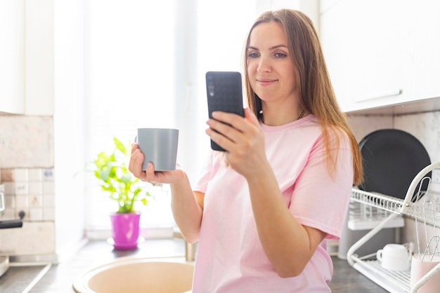 Beautiful young woman drinking coffee and using the phone in the kitchen