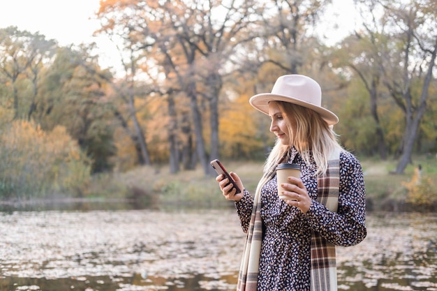 Beautiful young woman drinking coffee, using phone in nature in autumn park in fall.