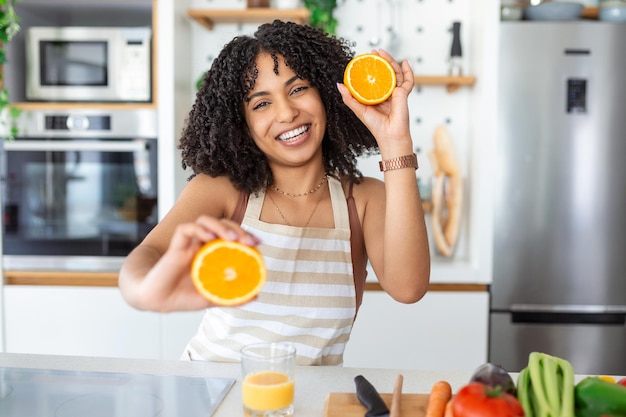 Beautiful young woman drinking fresh orange juice in kitchen Healthy diet Happy young woman with glass of juice and orange at table in kitchen