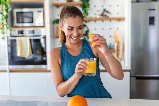 Beautiful young woman drinking fresh orange juice in kitchen Healthy diet Happy young woman with glass of juice and orange at table in kitchen