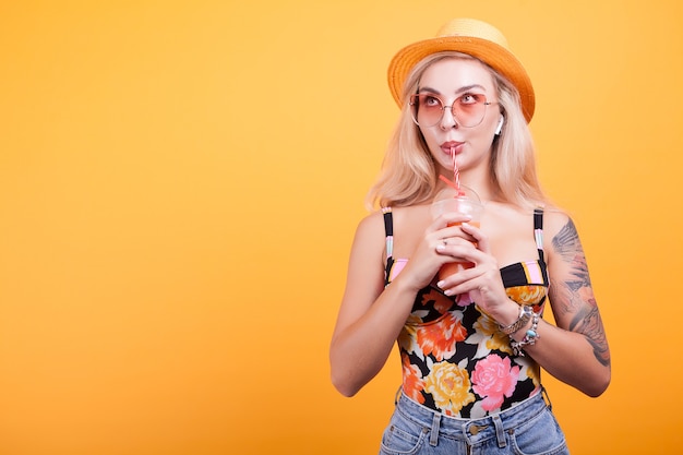 Beautiful young woman drinking fresh orange juice with sunglasses and hat in studio over yellow background