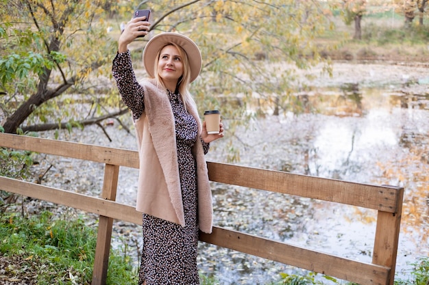 beautiful young woman drinks coffee, takes selfie on phone in nature in autumn park in fall