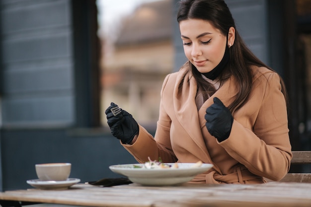 Beautiful young woman eating lunch in cafe outdoors on terrace. Female in black protective mask.