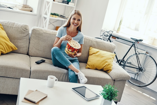 Beautiful young woman eating popcorn and smiling while watching TV on the sofa at home