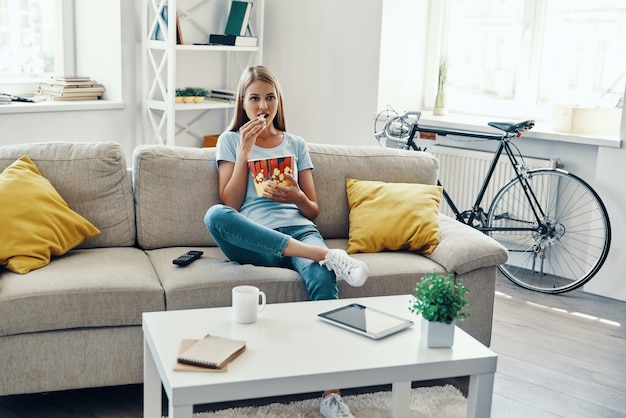 Beautiful young woman eating popcorn while watching TV on the sofa at home