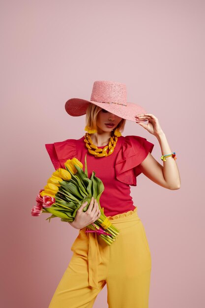 Photo beautiful young woman in elegant hat holding bunch of tulips against pink background