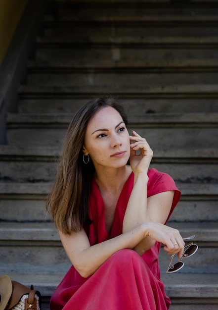 A beautiful young woman in an elegant red dress and a straw hat is sitting on the steps of a stone staircase overlooking the spring garden