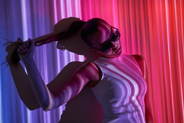 Beautiful young woman in eyeglasses adjusting hair while leaning at the wall with colorful lighting