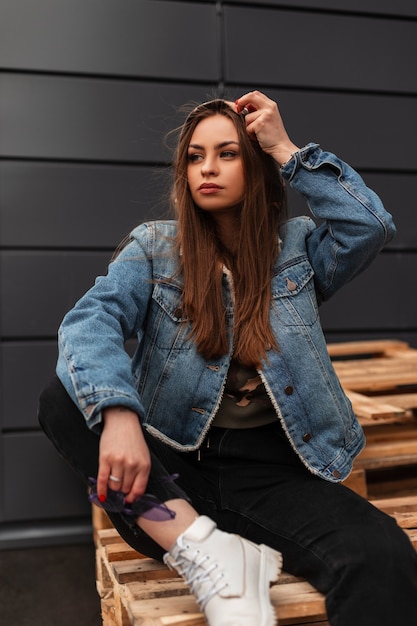 Beautiful young woman fashion model in fashionable denim clothes in stylish white shoes is resting on wooden planks near gray building. Urban pretty modern trendy girl sits on wooden pallets outdoors