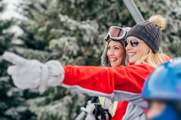 Photo beautiful young woman friends enjoying in winter vacations. they driving on ski lift and looking away with smile.