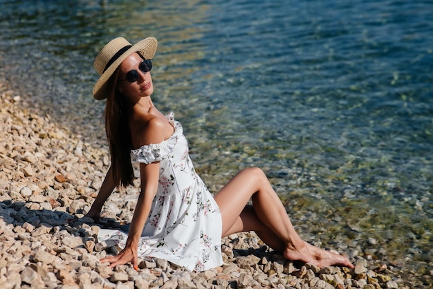 A beautiful young woman in a hat, glasses and a light dress is sitting on the ocean shore against the background of huge rocks on a sunny day. Tourism and tourist trips.