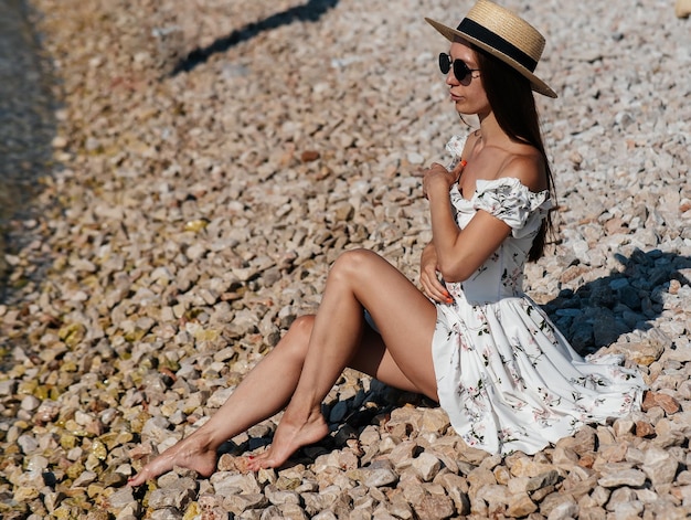 A beautiful young woman in a hat glasses and a light dress is sitting with her back to the ocean against the background of huge rocks on a sunny day Tourism and tourist trips