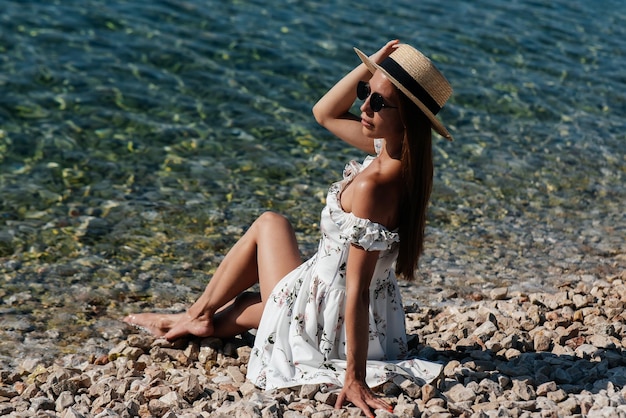 A beautiful young woman in a hat glasses and a light dress is sitting with her back to the ocean against the background of huge rocks on a sunny day Tourism and tourist trips