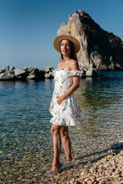 A beautiful young woman in a hat glasses and a light dress is walking along the ocean shore against the background of huge rocks on a sunny day Tourism and tourist trips