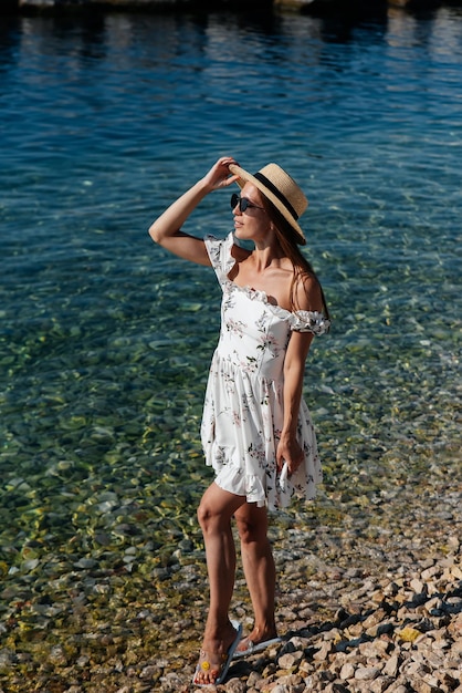 A beautiful young woman in a hat glasses and a light dress is walking along the ocean shore against the background of huge rocks on a sunny day Tourism and tourist trips