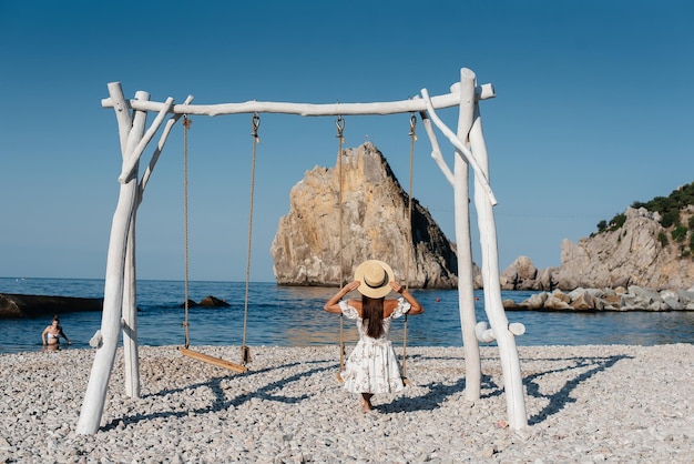 A beautiful young woman in a hat and a light dress is riding on a swing on the ocean.