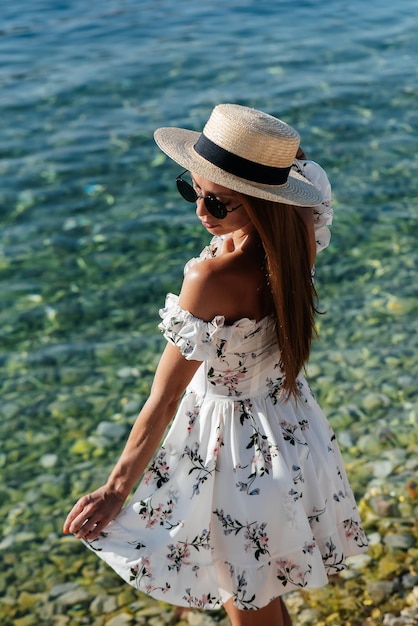 A beautiful young woman in a hat and a light dress with her back is walking along the ocean shore against the background of huge rocks on a sunny day Tourism and vacation travel