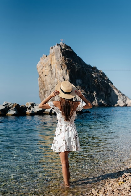 A beautiful young woman in a hat and a light dress with her back is walking along the ocean shore against the background of huge rocks on a sunny day Tourism and vacation travel