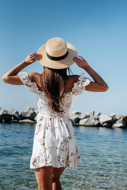 A beautiful young woman in a hat and a light dress with her back is walking along the ocean shore against the background of huge rocks on a sunny day Tourism and vacation travel