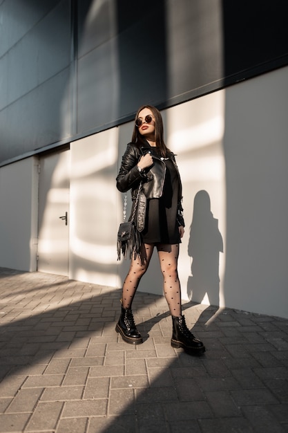 Beautiful young woman hipster in black leather fashionable clothes with stylish sunglasses and boots walks on the street near a modern building at sunlight