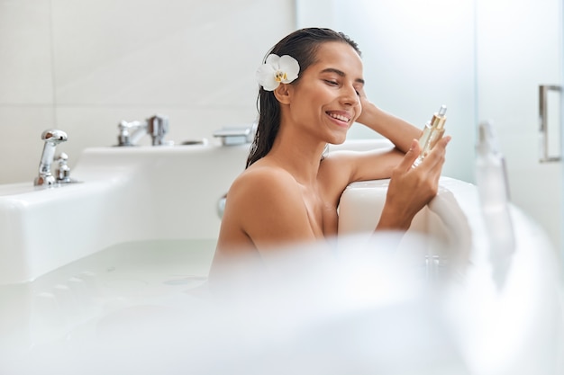 Beautiful young woman holding bottle of face serum while taking bath