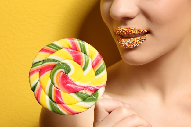 Beautiful young woman holding lollipop near lips covered with sprinkles on color background closeup