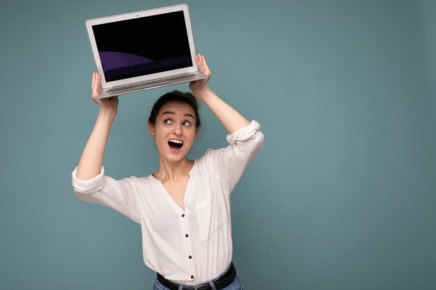 Beautiful young woman holding netbook computer looking up and saying wow wearing white shirt