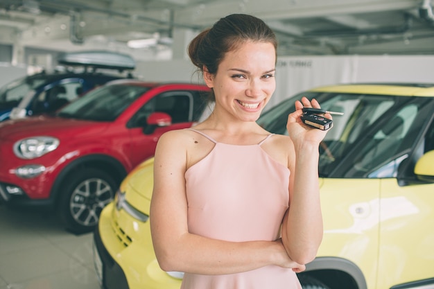 Beautiful young woman is holding a key in car dealership. Auto business, car sale, - happy female model in auto show or salon.