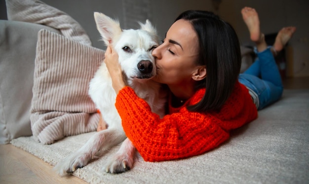 Beautiful young woman is hugging and kissing her big white cute dog while lying on the carpet on home floor