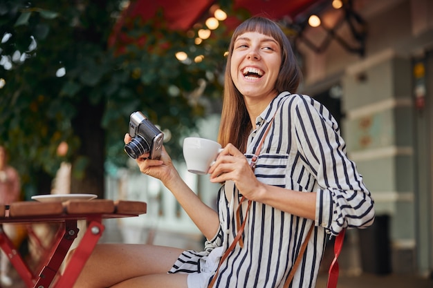 Beautiful young woman is laughing while holding camera and cup of coffee