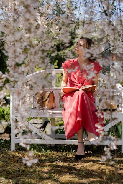 A beautiful young woman is sitting on an elegant bench in a spring garden under a cherry blossom and reading a book