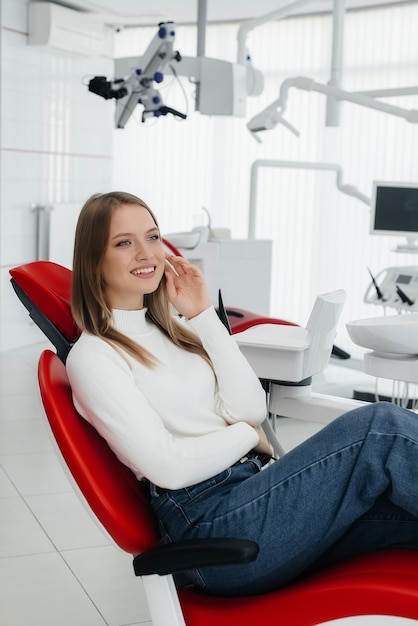 A beautiful young woman is sitting on a red dental chair in modern white dentistry and smiling Dental prosthetics treatment and teeth whitening