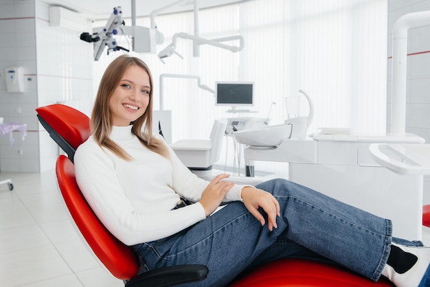 A beautiful young woman is sitting on a red dental chair in modern white dentistry and smiling Dental prosthetics treatment and teeth whitening