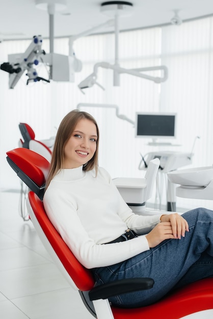 A beautiful young woman is sitting on a red dental chair in modern white dentistry and smiling Dental prosthetics treatment and teeth whitening