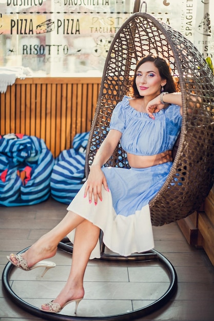 A beautiful young woman is sitting in a wicker hanging chair on the summer terrace of a city cafe looking at the camera and smiling