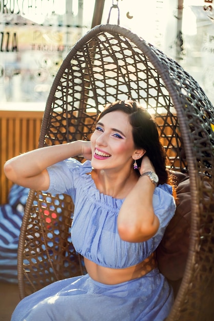 A beautiful young woman is sitting in a wicker hanging chair on the summer terrace of a city cafe looking at the camera and smiling