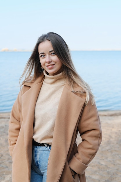 A beautiful young woman is standing on the beach against the backdrop of blue water smiling and looking at the camera Slow living Enjoying the little things Lykke concept