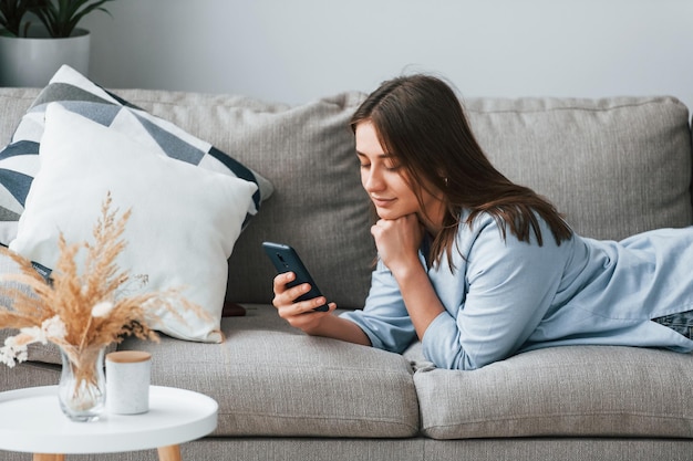 Beautiful young woman in jeans and blue shirt lying down at sofa with phone in hand