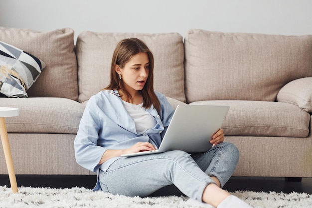 Beautiful young woman in jeans and blue shirt sits on the floor with laptop indoors at home