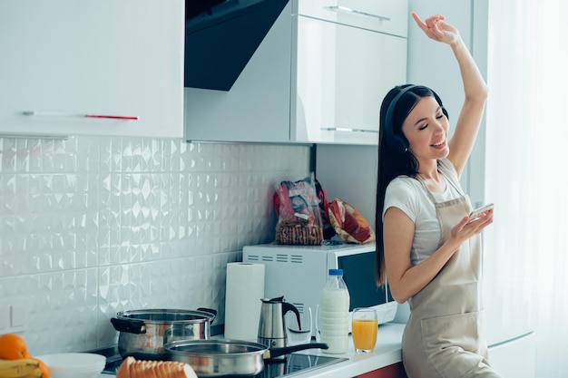 Beautiful young woman in the kitchen putting hand up while holding a smartphone and enjoying the music