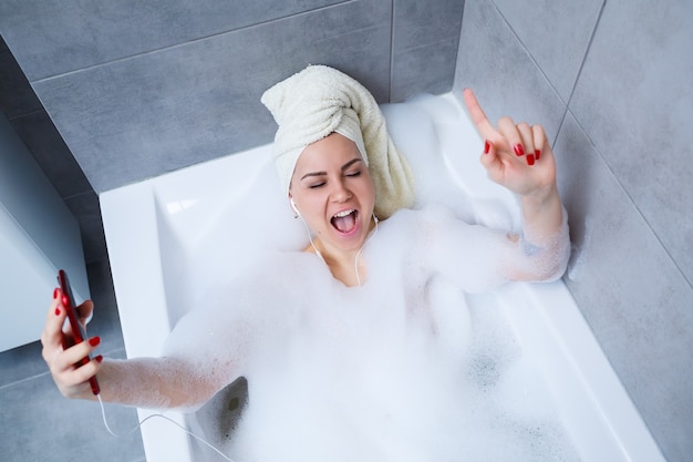 Beautiful young woman listening to music and singing while taking a bath at home.  Relax after a hard day. Spa relaxing procedure.