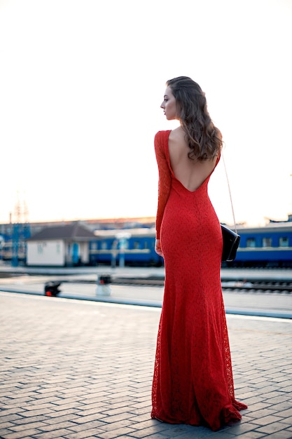 Beautiful young woman in a long red dress stands on the platform of the station near the railway