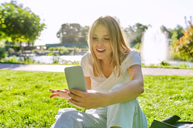 Beautiful young woman making video call on smartphone, female sitting on grass in park, rest online communication with friends, student talking, blogger blogging blog vlog