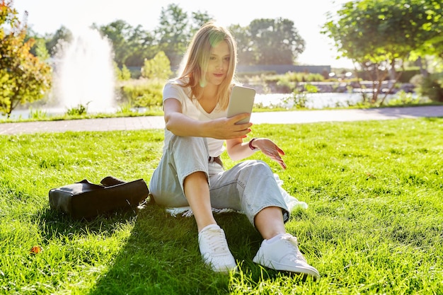 Beautiful young woman making video call on smartphone, female sitting on grass in park, rest online communication with friends, student talking, blogger blogging blog vlog