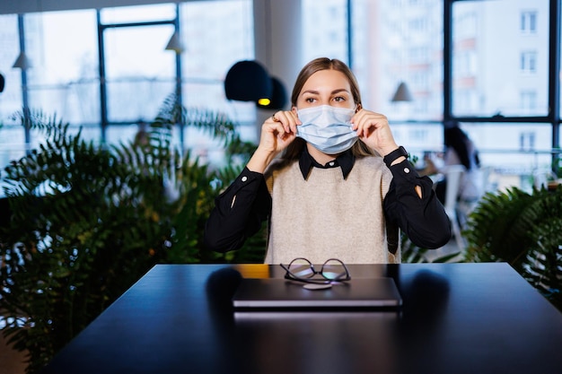 Beautiful young woman in a mask working in a cafe with a laptop during the coronavirus pandemic.