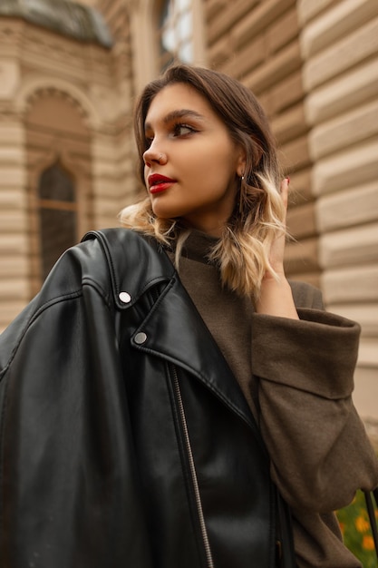 Beautiful young woman model with red lips in fashionable black leather jacket and green sweater walks in the city on an autumn day